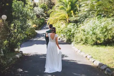 Un couple de mariés avance sur une allée bordée de verdure luxuriante, baignée de lumière naturelle, illustrant l’atmosphère romantique et élégante d’un domaine de mariage au Pays Basque.